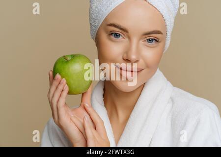 Femme aux yeux bleus avec un maquillage minimal bien entretenu teint frais la peau propre, vêtue d'un manteau blanc, retient la pomme verte isolée sur fond marron Banque D'Images