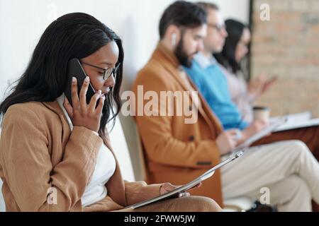 Vue latérale d'une jeune femme noire sérieuse assise sur une chaise dans le couloir et parler par téléphone en attendant le travail entretien Banque D'Images