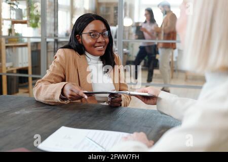 Jeune candidat noir souriant en lunettes assis à table et de donner le presse-papiers au recruteur tout en présentant des informations à son sujet expérience Banque D'Images