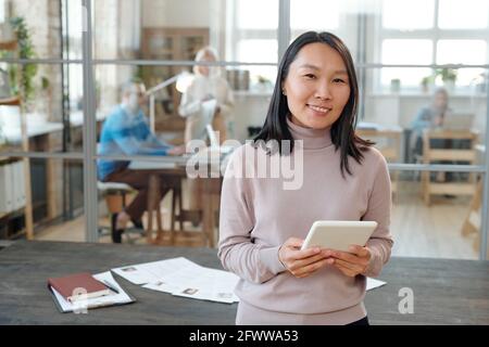Portrait d'un jeune responsable RH asiatique souriant avec une tablette debout contre table en bois avec curriculum vitae des candidats Banque D'Images