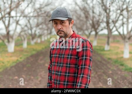 Portrait d'un beau fermier confiant avec casquette de baseball et chemise en flanelle debout dans le verger de fruits de noyer et regardant la caméra, mise au point sélective Banque D'Images