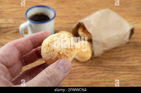 Main tenant un petit pain au fromage brésilien typique devant des pains non concentrés et une tasse de café. Banque D'Images