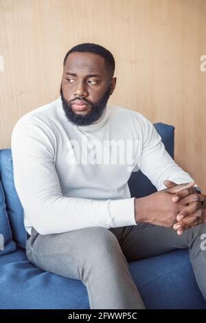 Homme afro-américain sérieux et réfléchi, assis sur un canapé de côté Banque D'Images