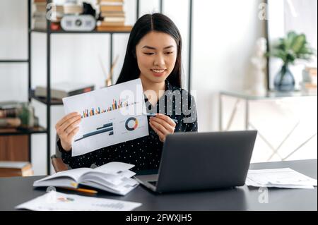 Jeune adulte sympathique souriant Femme asiatique confiante en chemise formelle, assise à son bureau, analyse de rapport, montrant le graphique financier à ses collègues pendant la communication par vidéoconférence avec Banque D'Images