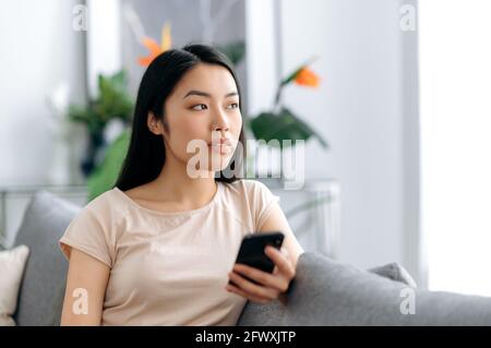 La jeune femme asiatique pensive utilise son téléphone mobile tout en étant assise sur le canapé dans des vêtements décontractés, navigue sur Internet et les réseaux sociaux, envoie des SMS avec des amis ou de la famille, regarde soigneusement Banque D'Images