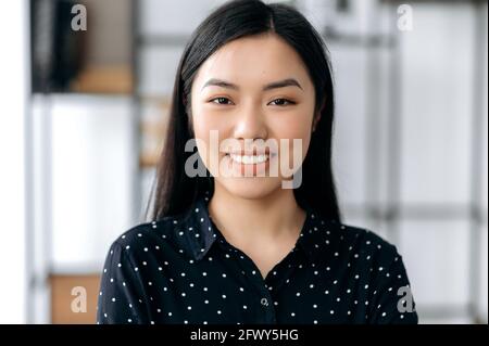 Portrait en gros plan d'une jolie fille asiatique confiante et heureuse. La jeune femme brune japonaise, portant une chemise noire élégante, se tenir sur un arrière-plan de bureau flou, regarde directement l'appareil photo, sourit amical Banque D'Images