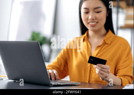 Achats en ligne. Heureux flou belle jeune femme asiatique brune assis à la table, utilise l'ordinateur portable et la carte de crédit pour payer pour les achats et la livraison en ligne, saisit des données, regarde la carte, sourit Banque D'Images