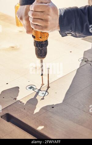 Un employé en blouson gris assemble un nouveau tour de table en bois vis avec tournevis électrique à l'extérieur, le jour ensoleillé, gros plan extrême Banque D'Images