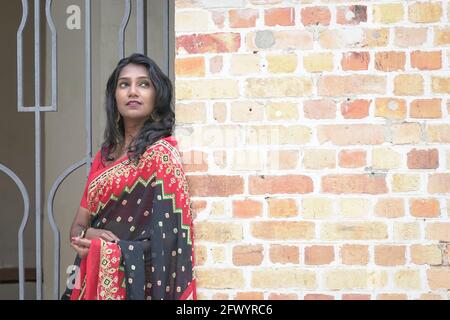 Portrait d'une femme indienne belle et souriante portant une saree rouge. Debout à côté du mur de briques, cadre extérieur. Banque D'Images