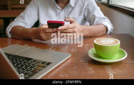 Homme lisant un téléphone avec ordinateur et une tasse de café sur une table. Vue rapprochée de la saisie de doigts sur le téléphone. Banque D'Images