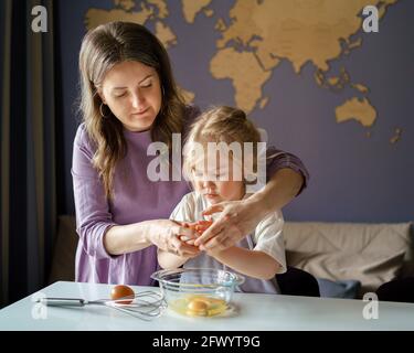Impliquer les enfants dans le processus de cuisson. Jeune maman montrant une petite fille mignonne comment casser des œufs frais dans un bol pour la tarte ou l'omelette tout en préparant les aliments ensemble à la maison. Mère enseignant enfant à cuisiner Banque D'Images