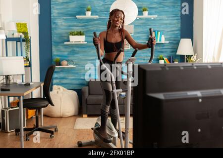 Active fitness stong femme noire courant sur machine elliptique, dans le salon à la maison regardant le tutoriel d'un instructeur en ligne. Personne sportive suivant le programme de cross. Banque D'Images