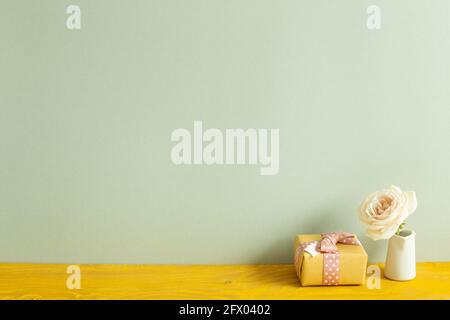 Boîte cadeau jaune et vase de fleur rose blanche sur table en bois. Fond vert kaki Banque D'Images