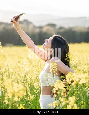 Vue latérale sourire mince brunette en blanc de l'été de vêtements de prendre sélectionnez votre smartphone tout en vous tenant sur un champ de colza en pleine floraison temps ensoleillé Banque D'Images