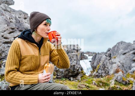 Jeune femme aux cheveux teints portant des vêtements chauds et ayant boisson chaude avec sandwich tout en étant assis sur le rocher dans la prairie Des sommets de l'Europe Banque D'Images
