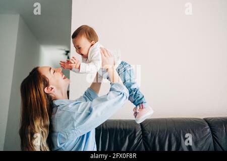 Vue de côté bonne jeune maman dans une chemise décontractée élevant mignon rire bébé sur les mains tout en passant du temps ensemble dans la lumière séjour Banque D'Images