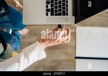Vue de dessus de la récolte photographe femelle méconnaissable insérant une carte mémoire de l'appareil photo dans l'ordinateur portable assis à une table en verre Banque D'Images