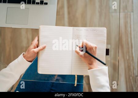 De dessus de récolte anonyme étudiante avec cheveux bouclés dans des vêtements décontractés, assis à une table en verre et prenant des notes dans le copybook lors de la préparation de Banque D'Images