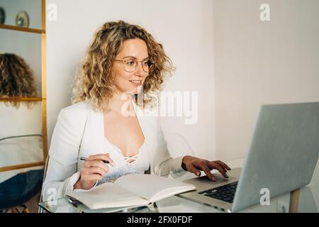 Jeune femme freelance avec cheveux blond courbé dans des vêtements décontractés et des lunettes de vue prenant des notes dans le planificateur et regardant loin pendant travailler à distance à l'aide de l Banque D'Images
