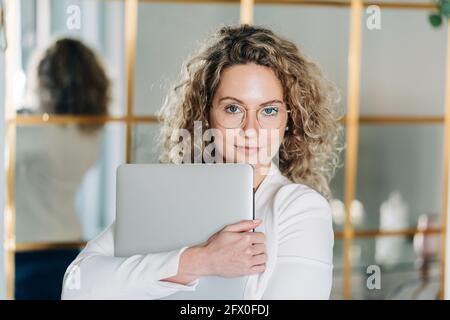 Jeune entrepreneur auto-assuré avec cheveux blonds bouclés dedans blouse blanche et lunettes de vue regardant l'appareil photo tout en étant debout espace de travail moderne avec Banque D'Images