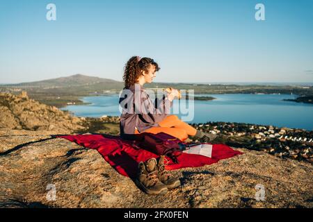 Vue latérale d'une jeune femme heureuse dans des vêtements décontractés manger un sandwich tout en étant assis sur une couverture sur une colline rocheuse mer contre ciel bleu sans nuages Banque D'Images