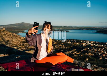 Vue latérale d'une jeune femme heureuse dans des vêtements décontractés manger un sandwich et prendre un selfie avec un téléphone portable tout en étant assis sur une couverture sur une colline rocheuse Banque D'Images