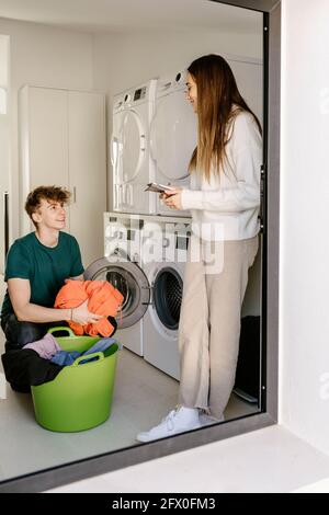 Joyeux jeune gars dans une tenue décontractée chargement des vêtements dans le lavage machine et sourire tout en communiquant avec une petite amie positive à l'aide d'un smartphone en mois Banque D'Images