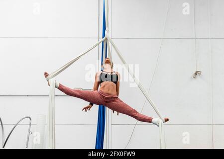 Femme sportive sportive et agile, non reconnaissable, dans des vêtements d'activité qui effectuent des fractionnements sur les silks aériens pendant l'entraînement en studio de gymnastique Banque D'Images