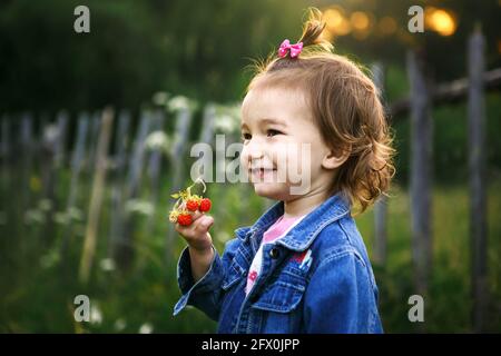 Une petite fille de 2 ans dans le village tient une fraise mûre dans ses mains et sourit sly. Vitamines, heure d'été. Journée internationale des enfants Banque D'Images