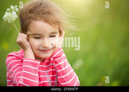 Une petite fille dans un T-shirt rayé corail sur un fond vert dans un champ tient son visage dans ses mains et sourit sly. Journée des enfants, enfant heureux, env Banque D'Images