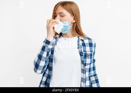 Malade jeune femme, portant un masque médical de protection soufflant son nez, elle a une fièvre, elle a attrapé un froid, éternuant sur un fond blanc isolé Banque D'Images