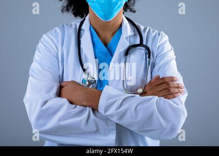 Section médiane de la femme médecin portant un masque facial debout avec bras croisés sur fond gris Banque D'Images