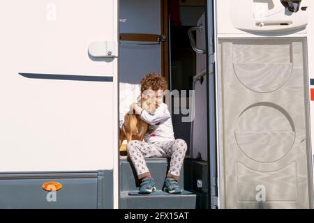 petit garçon aux cheveux bouclés souriant assis dans la porte de la maison de moteur, tenant tendement son animal de compagnie. Banque D'Images