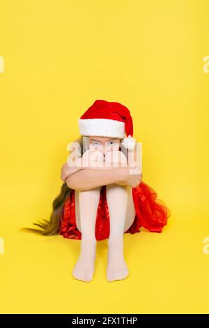 Triste jeune fille en robe rouge et le chapeau rouge du Père Noël est assis et embrasse ses genoux. Fille dans chapeau de Santa isolé sur fond jaune. Banque D'Images