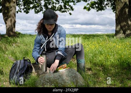 La fille a le déjeuner dans la nature. Thermos, café, fromage et sandwich à l'ardoise. Soleil, arbres, herbe verte, fleurs, ciel bleu et nuages de pluie. Par Banque D'Images