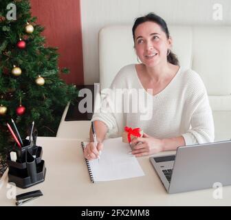 Jeune femme en chandail blanc écrit des notes au bureau à la maison, tient la boîte cadeau dans la main, prépare pour noël.Vente de vacances d'hiver Banque D'Images