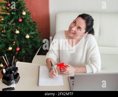 Jeune femme en chandail blanc écrit des notes au bureau à la maison, tient la boîte cadeau dans la main, prépare pour noël.Vente de vacances d'hiver Banque D'Images