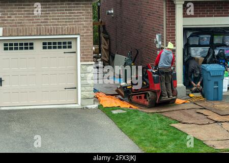 Un travailleur sur un petit tracteur livre des matériaux de construction une allée étroite entre les garages et un chantier de construction Banque D'Images