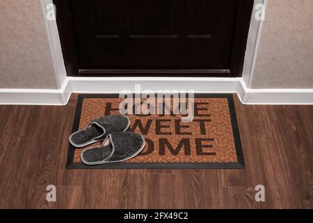 Paillasson avec texte Home Sweet home avant la porte sombre dans le hall. Chaussons à la maison sur le tapis. Vue de dessus du tapis sur le parquet. Banque D'Images