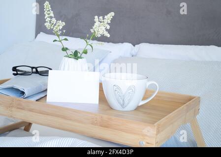 Petit-déjeuner dans le décor de maquette de papeterie de lit. Maquette de carte de vœux vierge, tasse de café, journal et verres sur plateau vase blanc avec petites fleurs. Val Banque D'Images