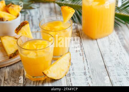 Délicieux jus d'ananas dans deux verres avec des tranches d'ananas. Cocktail d'ananas frais naturel et jus en verre Jug sur table blanche en bois avec palmier Banque D'Images