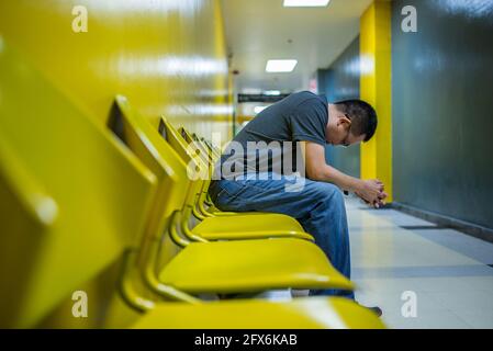 Homme asiatique déprimé, homme d'âge moyen assis seul sur une chaise jaune dans un bâtiment au niveau d'une allée, bras sur les genoux, vue latérale du corps, vue perspective. Banque D'Images
