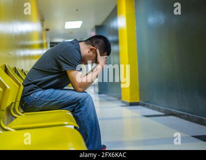 Homme asiatique déprimé, homme d'âge moyen assis seul sur une chaise jaune dans un bâtiment au niveau d'un chemin, mains tenant la tête, vue perspective, côté droit Banque D'Images