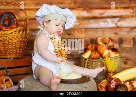 un petit boulanger, dans un chapeau de chef et un tablier avec un bouquet de bagels autour de son cou, prépare de la pâte pour cuire sur fond de produits de boulangerie. Banque D'Images