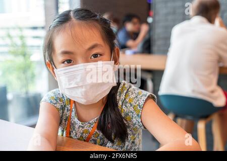 Fille asiatique à la table de nourriture de la cafétéria de nourriture au centre commercial de Bangkok, Thaïlande. Enfant fille portant un masque médical, regardant l'appareil photo, ba floue Banque D'Images