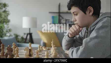 Un garçon confiant et intelligent jouant aux échecs à la maison, il pense et regarde les pièces disposées sur l'échiquier, le concept des jeux de stratégie Banque D'Images