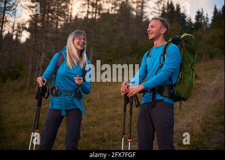 Couple de randonneurs s'amusant ensemble tout en marchant à l'extérieur sur fond de forêt. Couple de touristes souriant les uns aux autres tout en se retrouvant activement à la randonnée. Banque D'Images