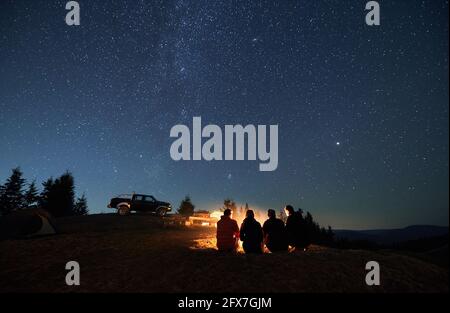 Le soir, ciel étoilé au-dessus de la vallée de la montagne avec voiture et randonneurs près du feu de camp. Groupe de voyageurs assis près d'un feu de joie sous un ciel bleu majestueux avec des étoiles. Concept de camping de nuit, de randonnée et de voyage. Banque D'Images