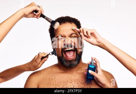 Mains de femmes se raser, appliquer le maquillage et le cologne à la personne barbu excitée. Homme criant avec joie tout en étant soigné par beaucoup de mains sur fond blanc Banque D'Images