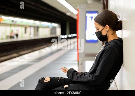 Adolescente assise sur un banc à la station de métro. Elle porte un masque de protection, un sweat-shirt et un Jean noir déchiré. Banque D'Images